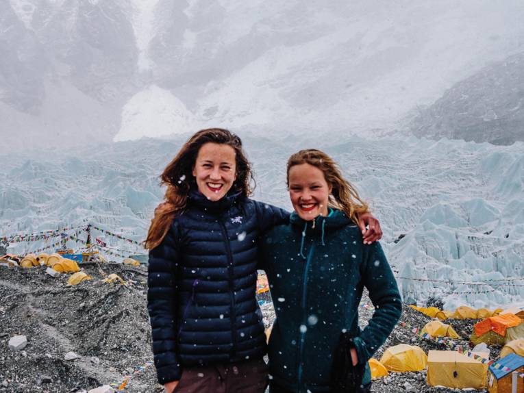 Auf dem Foto zu sehen sind zwei junge Frauen, die Arme um die Schultern gelegt. Sie stehen am Mount Everest im Basecamp. Es schneit